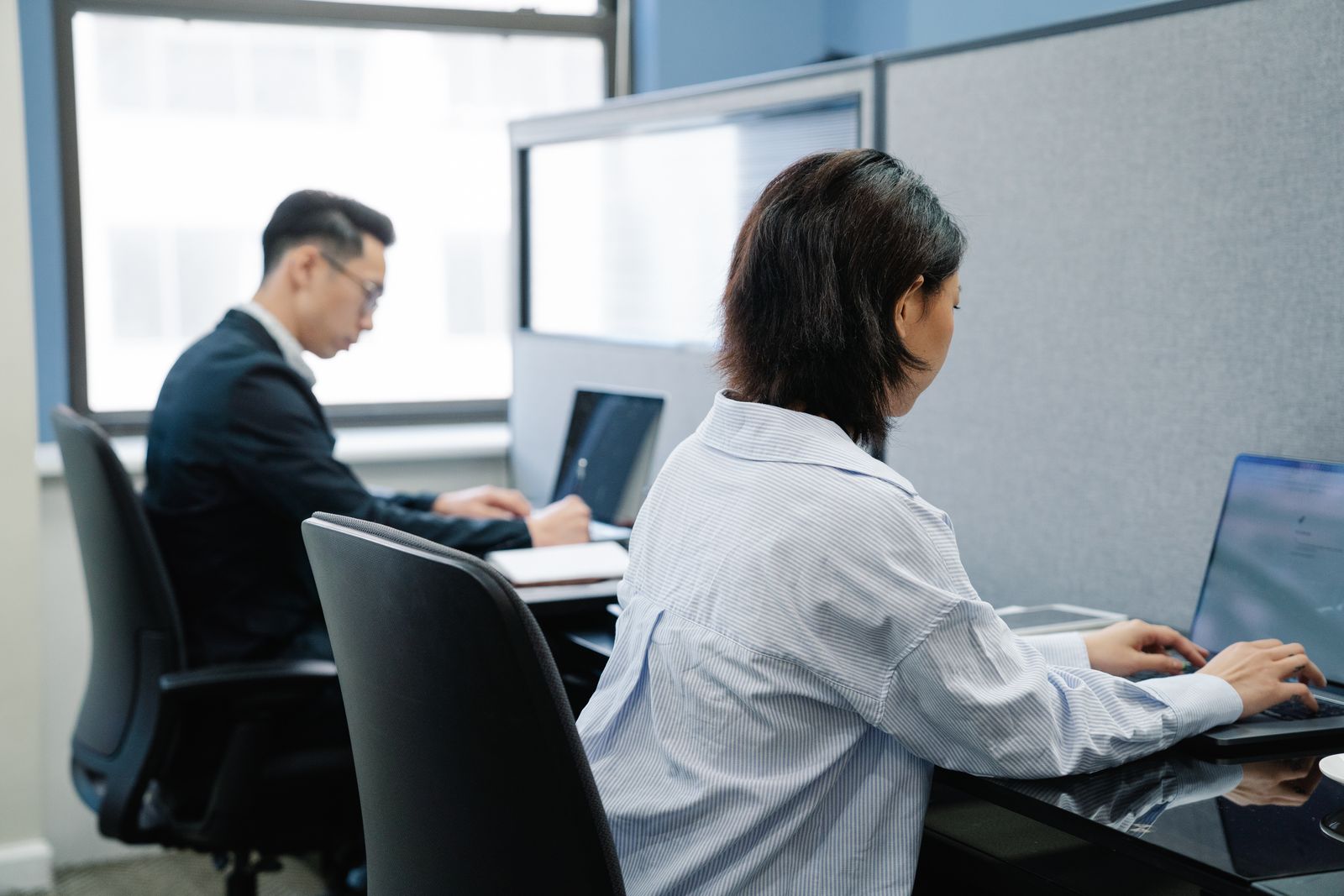Two office workers at laptops in separate cubicles beside a window